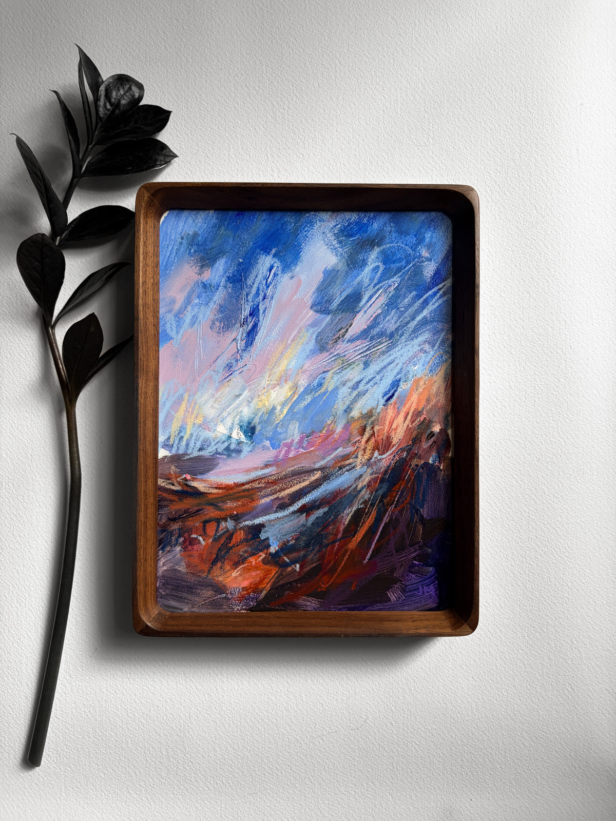 A framed abstract landscape painting featuring vibrant blue and brown brushstrokes, housed in a dark brown wooden frame and displayed on a light gray wall next to a black decorative branch.