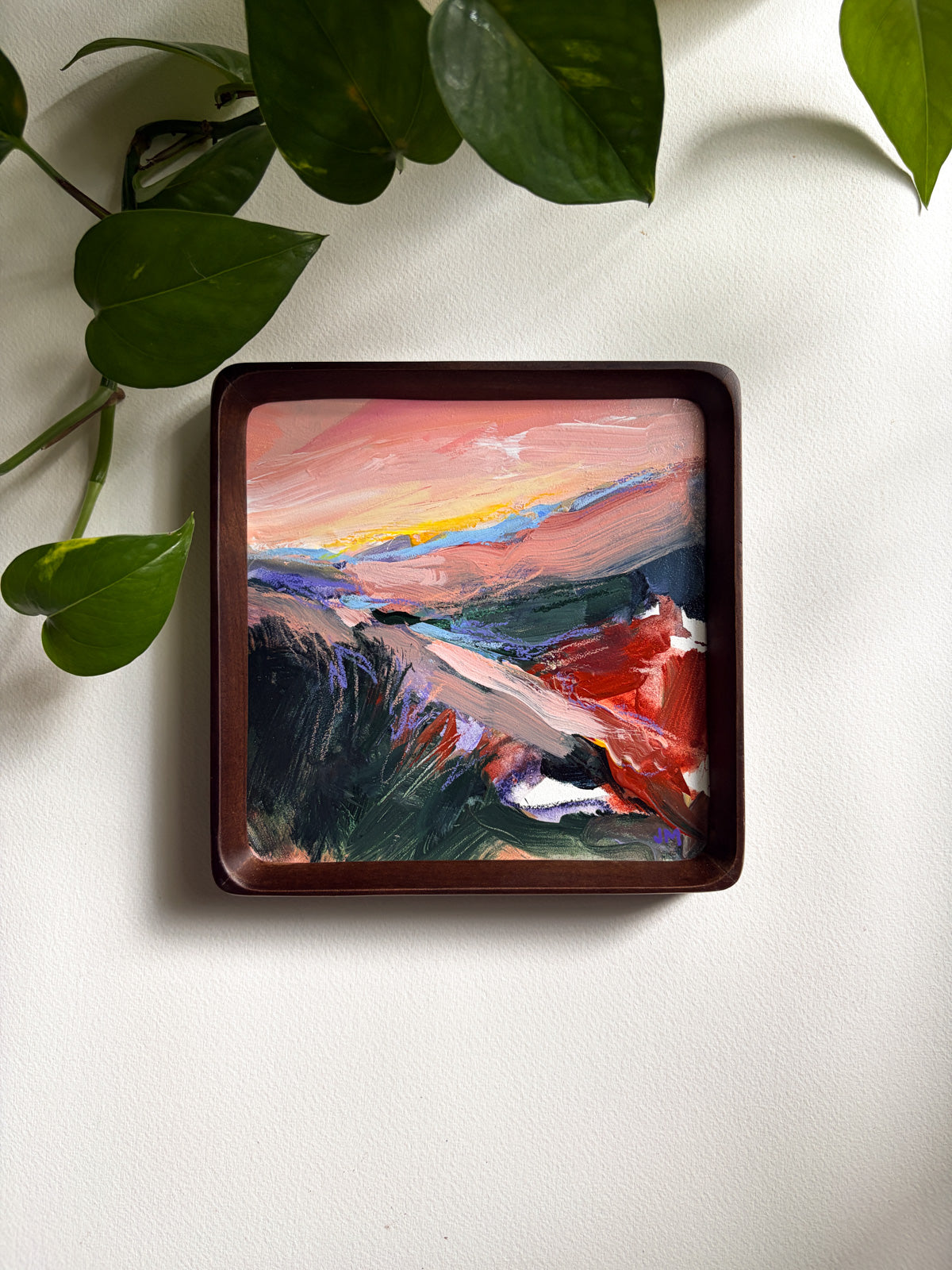A framed, colorful landscape painting, styled with green plant leaves on a white textured background.