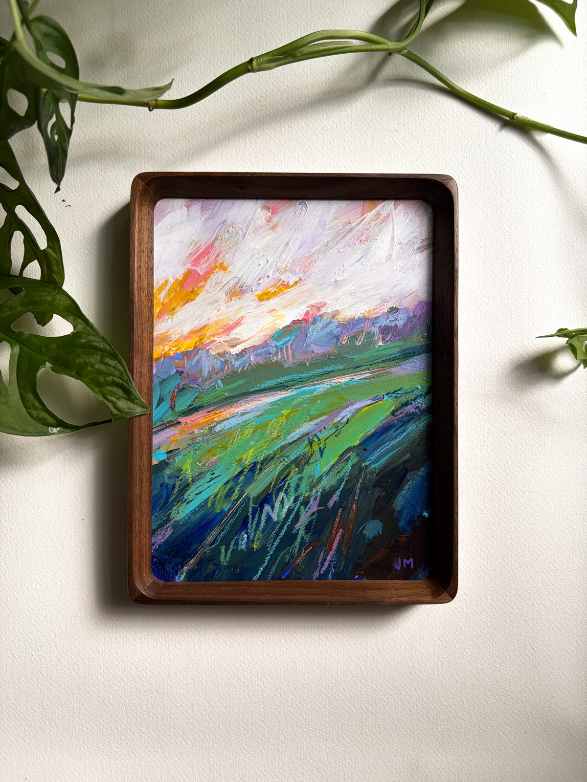 A framed abstract landscape painting with vibrant green, blue, purple, orange, and white brushstrokes, housed in a dark brown wooden frame and displayed on a textured off-white wall with green plant leaves in the foreground.