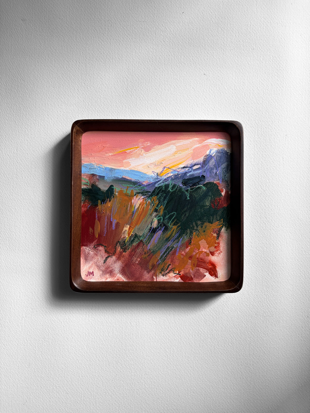 A square framed abstract landscape painting featuring pink, orange, green, and blue brushstrokes, mounted on a light gray textured wall.
