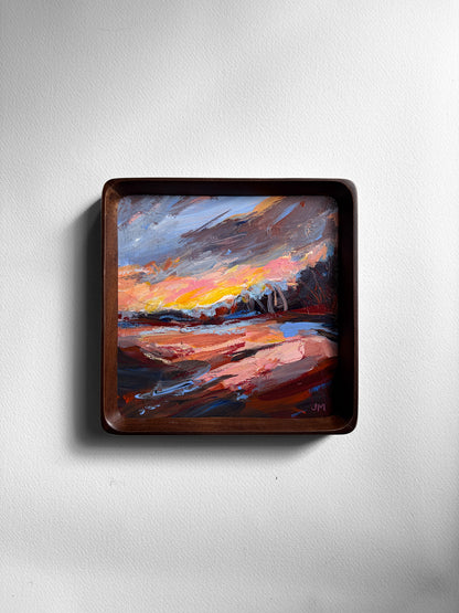A square framed landscape painting featuring a dramatic sunset or sunrise scene with swirling brushstrokes of orange, pink, blue, and brown, mounted on a light gray wall.