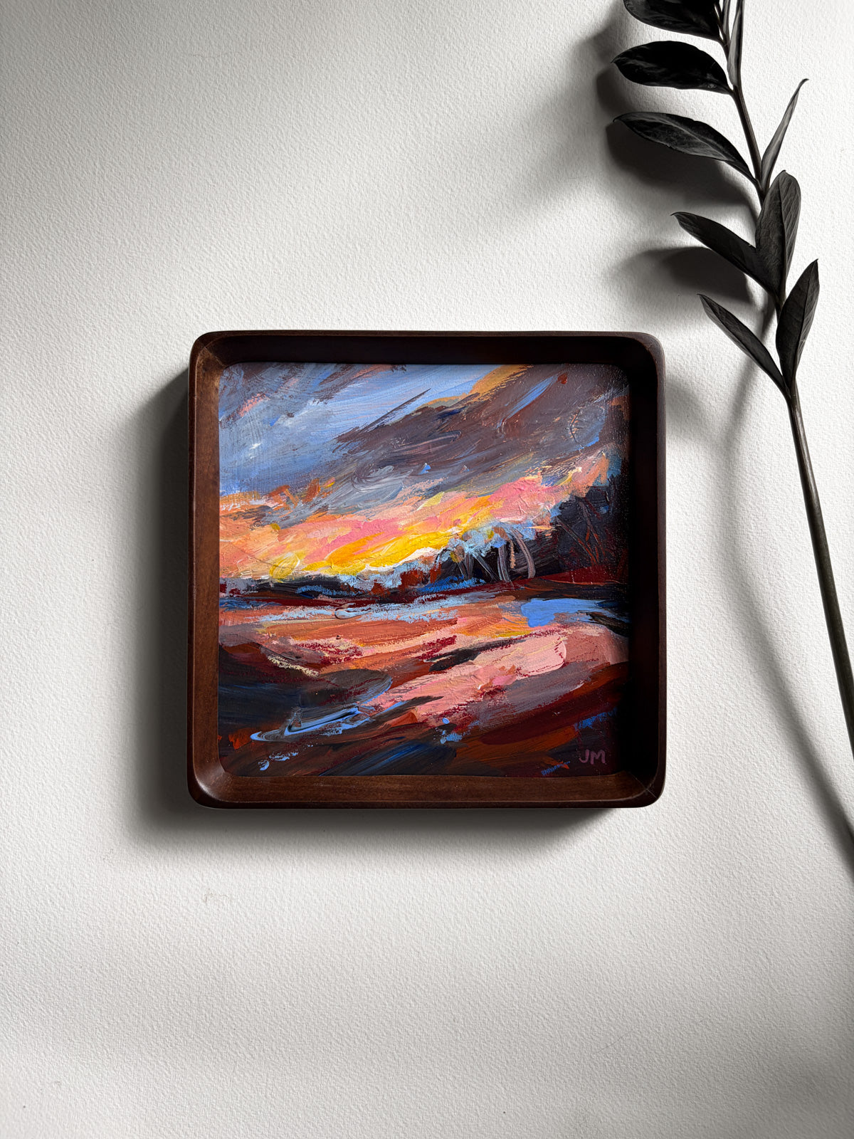 A square wooden-framed painting of a colorful sunset landscape, mounted on a light gray wall with a decorative plant leaf casting a shadow to the right.