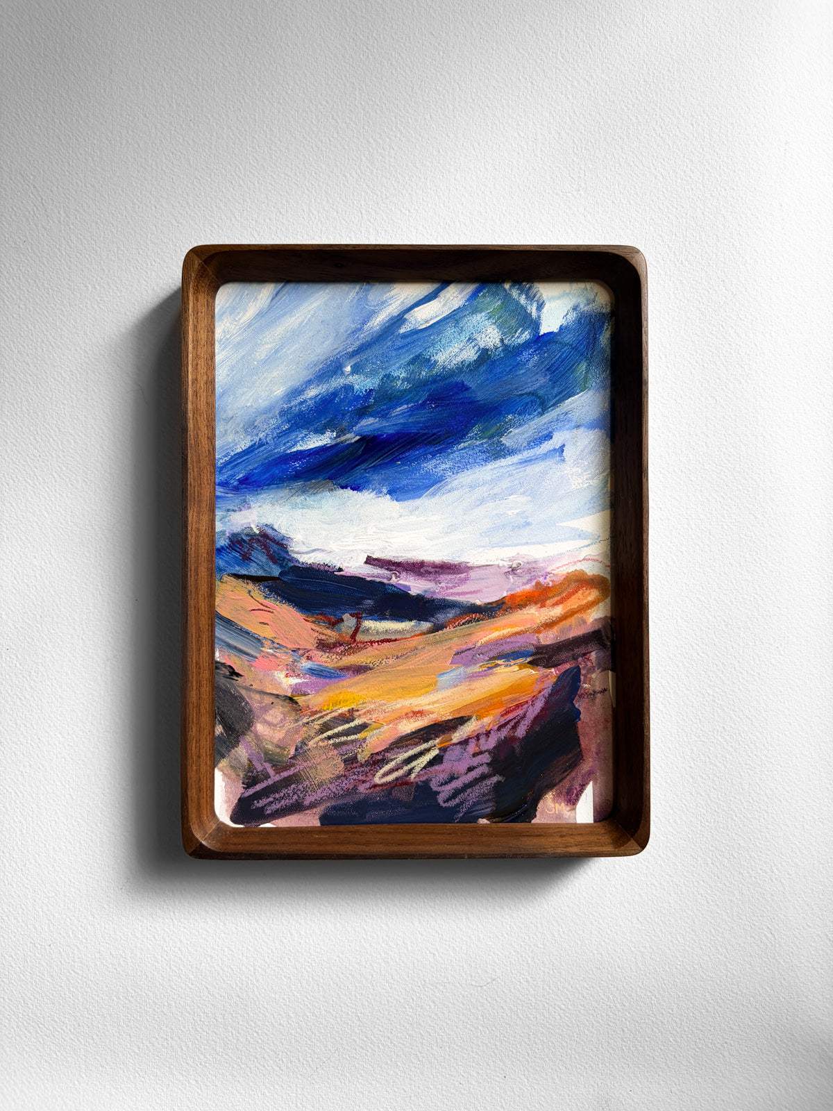 A framed abstract landscape painting featuring blue, purple, orange, and white brushstrokes, enclosed in a dark brown wooden frame and mounted on a light gray wall.