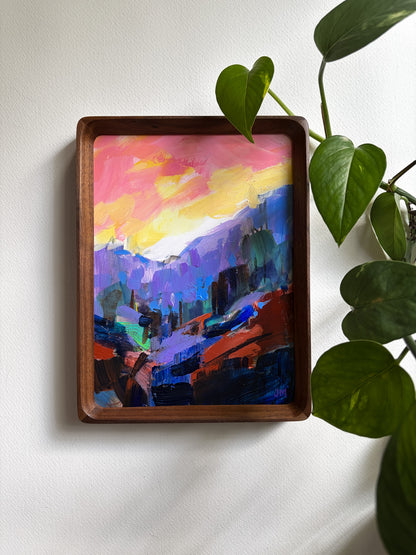 A framed colorful landscape painting hangs on a white wall, partially overlapped by green houseplant leaves. The artwork features a vivid sunset sky in pink, yellow, and orange above purple and blue mountains and valley.