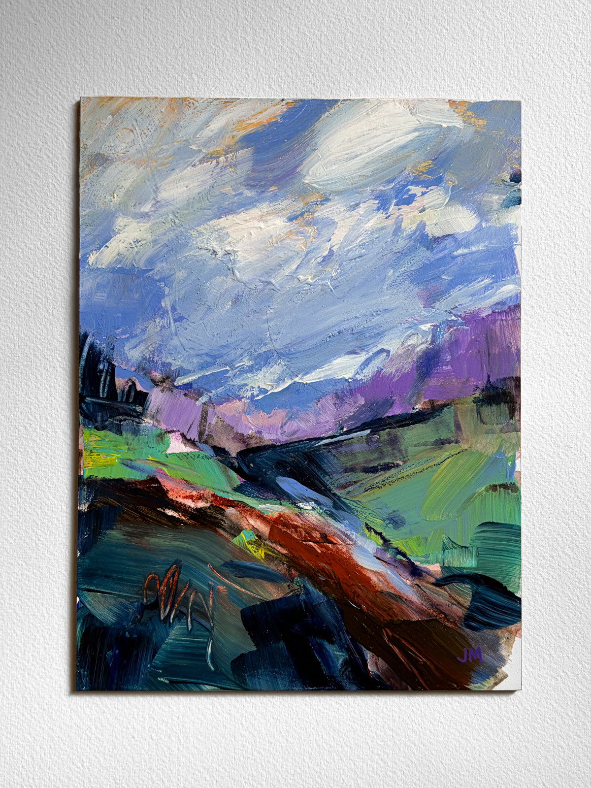 An abstract landscape painting on board, featuring a dramatic blue and white sky, purple mountains, green fields, and a pathway, displayed on a textured white wall.