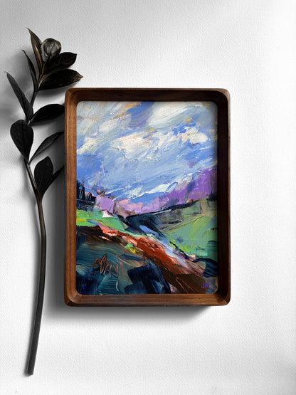 An abstract landscape painting in blue, purple, green, tones, styled on a white wall next to a dark decorative branch.