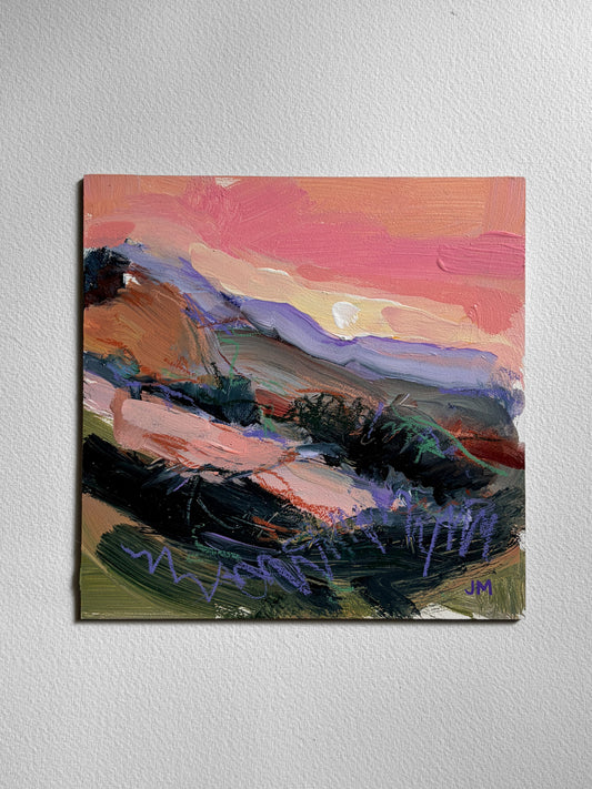 An abstract landscape painting on board, featuring a vibrant sunset sky in shades of pink, orange, and yellow, with layered purple and blue mountains and dark green foreground brushstrokes, displayed on a textured white wall.