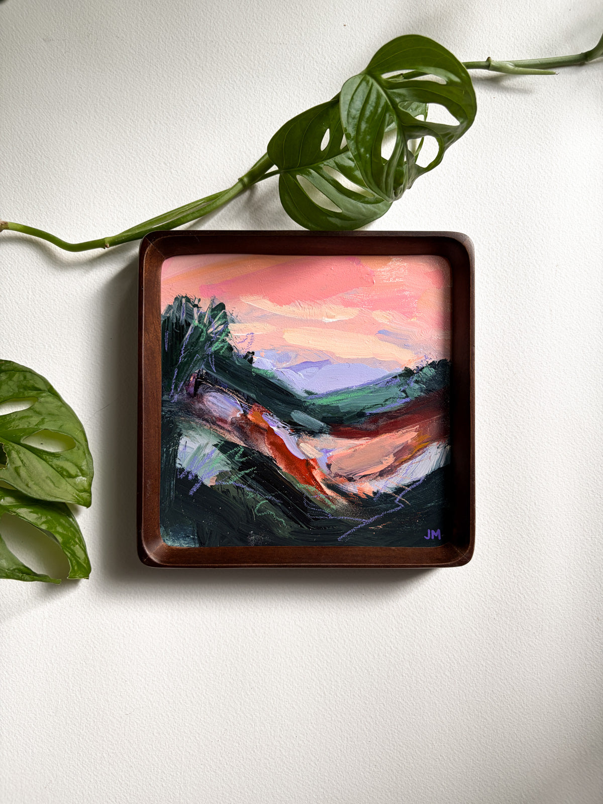 A square framed landscape painting with an abstract sunset scene, displayed on a white wall with green plant leaves in the corners.