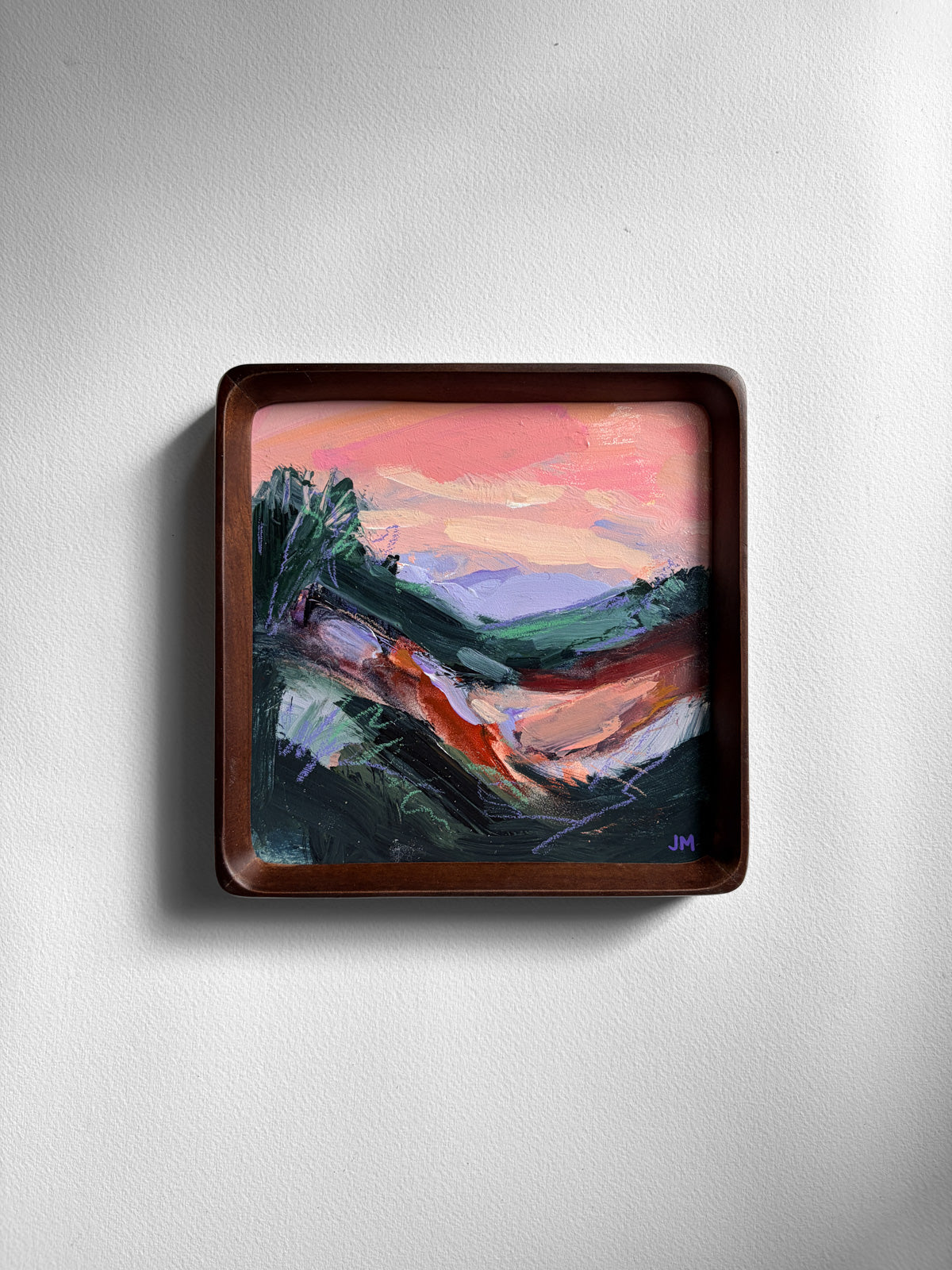 A square framed abstract landscape painting featuring a pink sky, violet hills, and dark hilly foreground, mounted on a light gray textured wall.