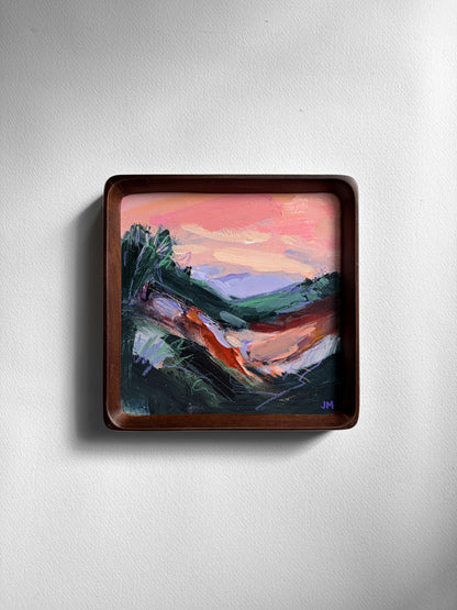 A square framed abstract landscape painting featuring a pink sky, violet hills, and dark hilly foreground, mounted on a light gray textured wall.