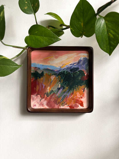 A square framed abstract landscape painting with a dark brown wooden frame, hanging on a white wall with green plant leaves framing the top of the image.