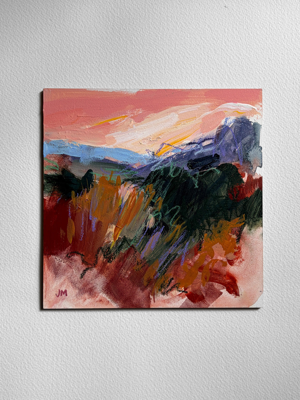 An abstract landscape painting on board, featuring a pink and orange sky, dark green and brown grasses, and blue mountains, displayed on a textured white wall.
