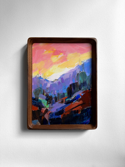 A framed abstract landscape painting featuring a colorful sunset sky in pink, yellow, and orange over purple mountains, housed in a dark brown wooden frame and mounted on a light gray wall.