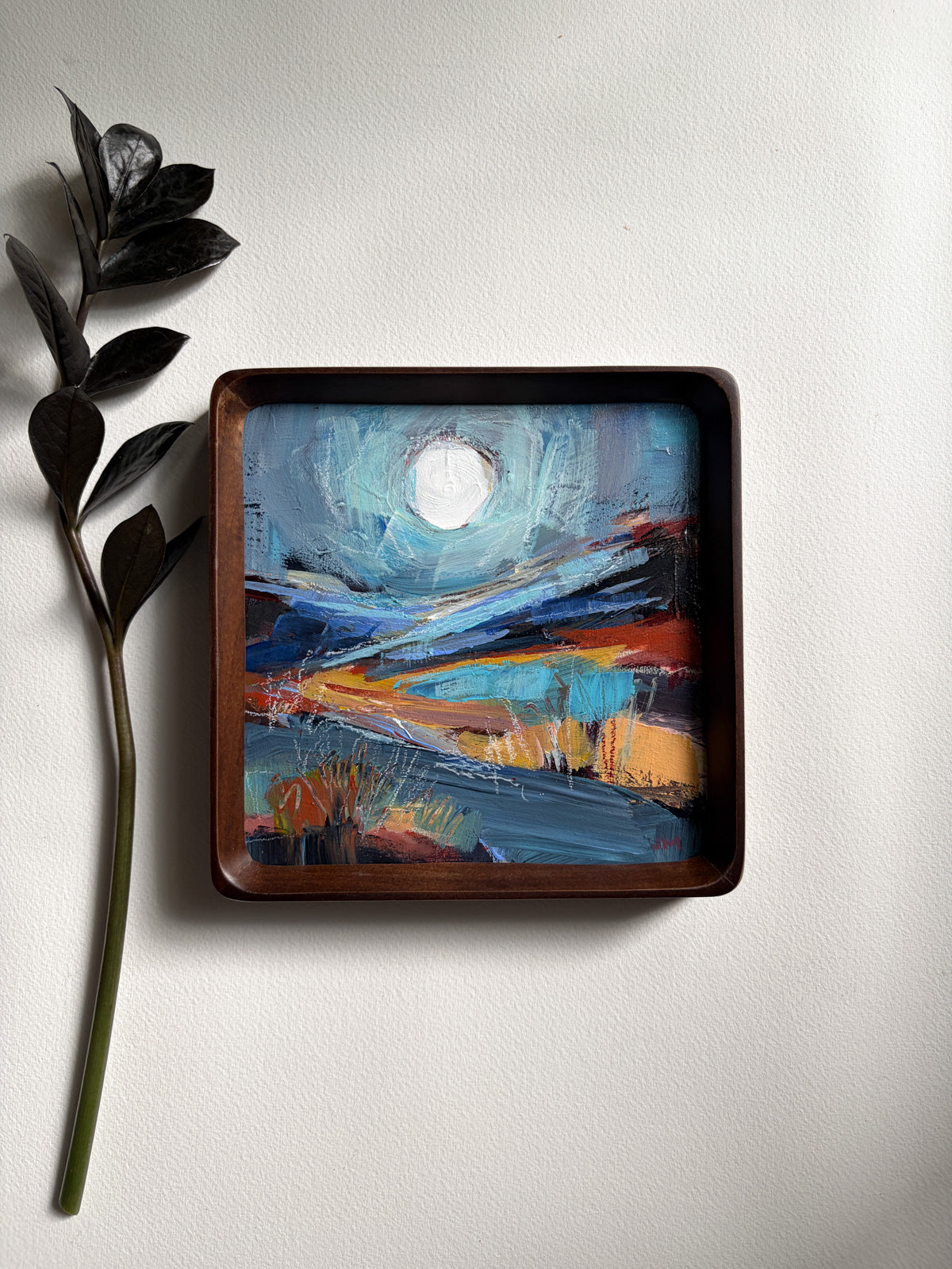 A square wooden frame containing an abstract landscape painting of a full moon, displayed on a light gray wall next to a dark decorative leaf stem.