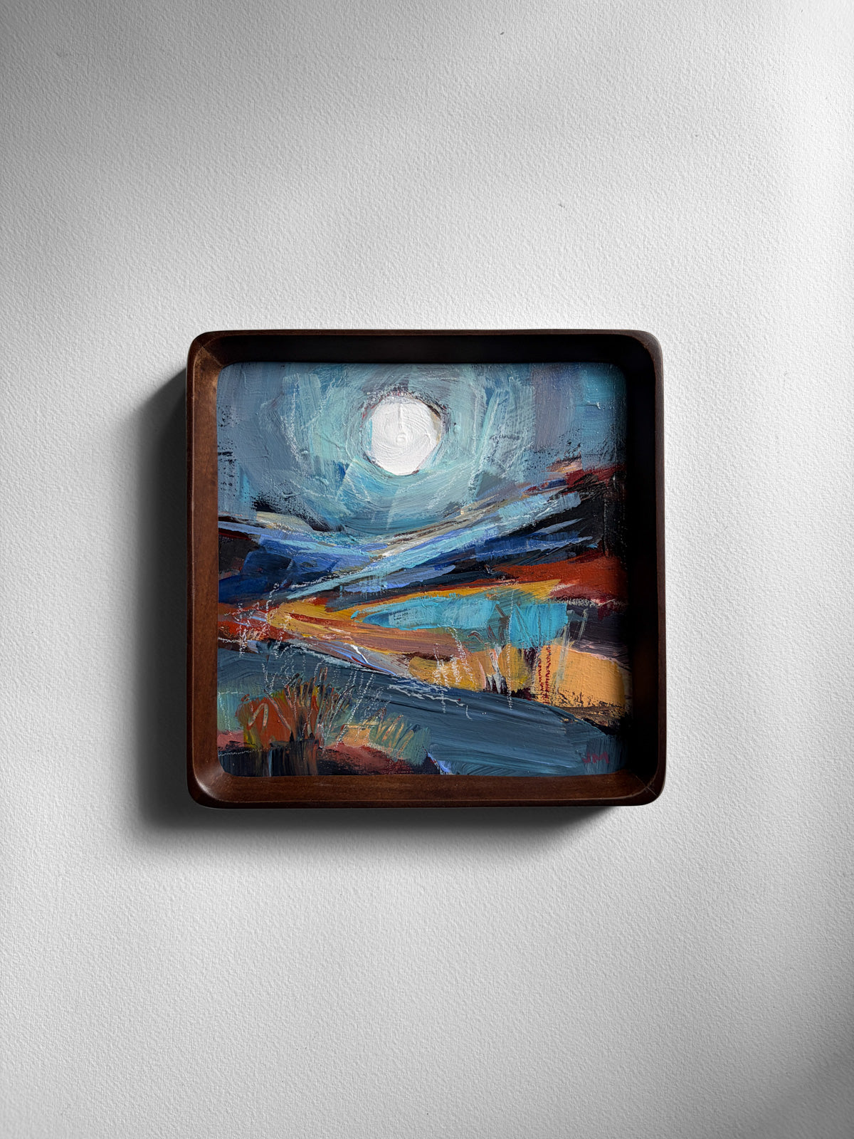 A square framed abstract landscape painting with a dark brown wooden frame, mounted on a light gray textured wall. The painting features a white moon in a turquoise sky.