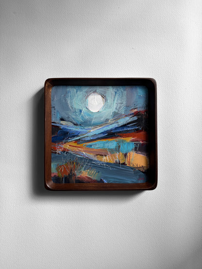 A square framed abstract landscape painting with a dark brown wooden frame, mounted on a light gray textured wall. The painting features a white moon in a turquoise sky.