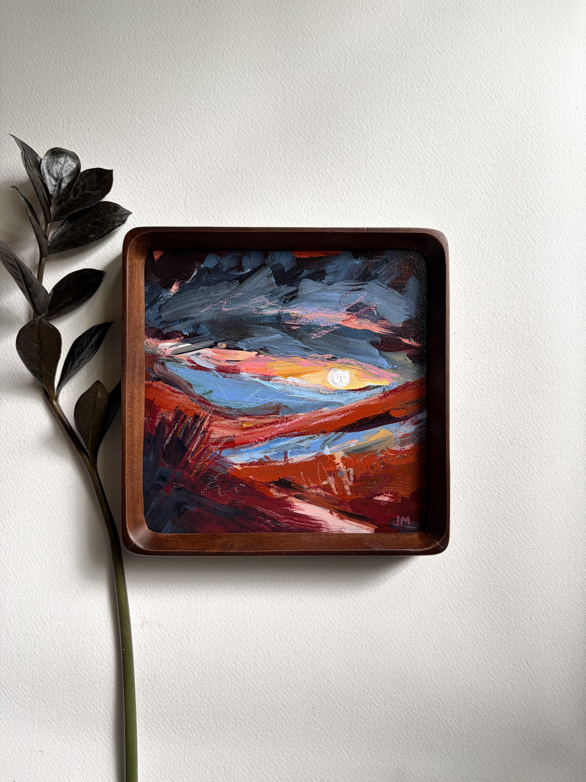 A square frame featuring an abstract landscape painting in red, orange, blue, and white, styled on a light gray wall next to a dark leafy stem.
