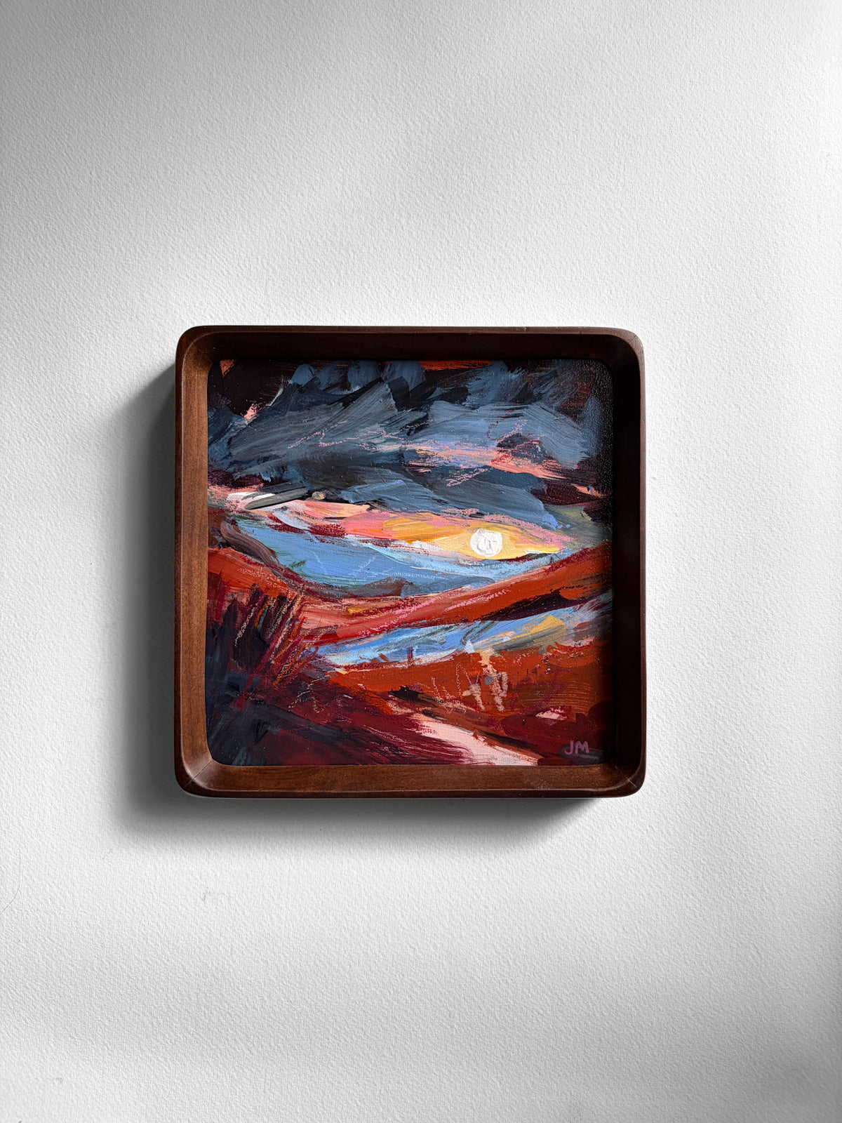 A square wooden frame with rounded corners, holding an abstract landscape painting in shades of red, blue, orange, and black, displayed on a light gray wall.