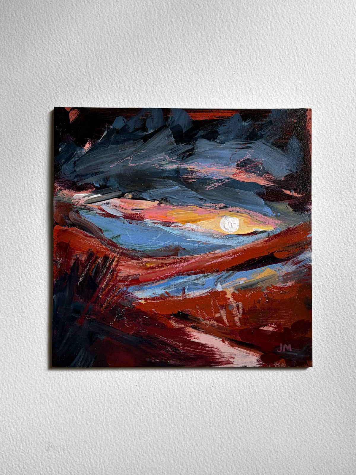 An abstract landscape painting on board, featuring expressive brushstrokes of deep red, dark blue, black, and orange, with a bright white moon near the center, displayed on a textured white wall.