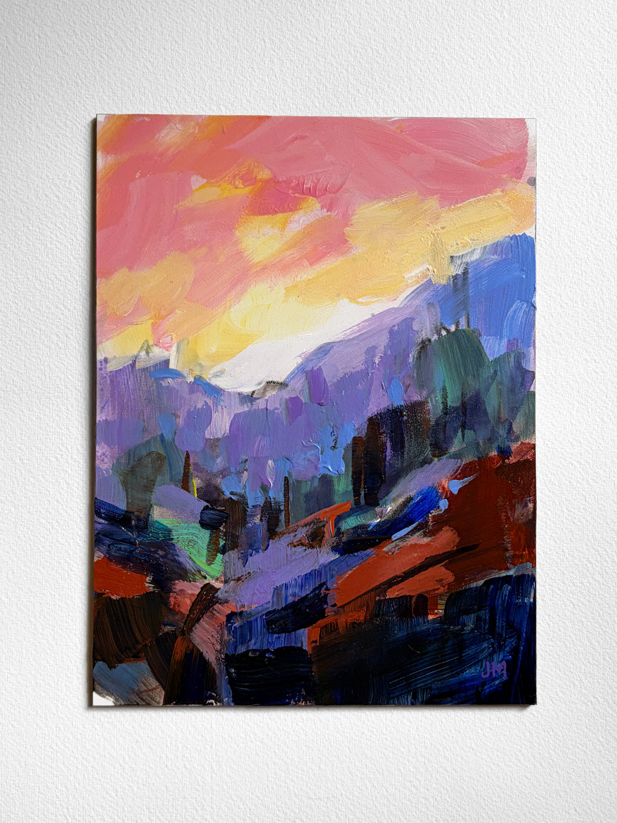 An abstract landscape painting on board, featuring a vibrant sunset sky in pink, yellow, and orange above purple mountains and dark blue foreground elements, displayed on a textured white wall.