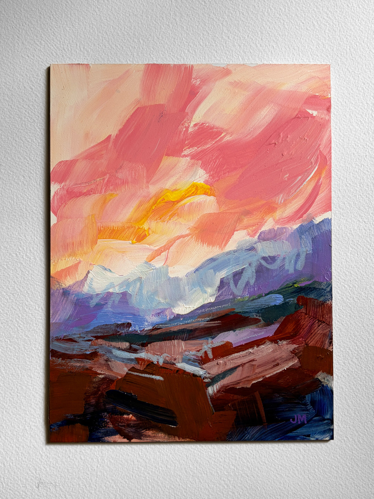 An abstract landscape painting on board featuring a vibrant pink and orange sky, purple mountains, and a dark brown foreground, displayed on a textured white wall.