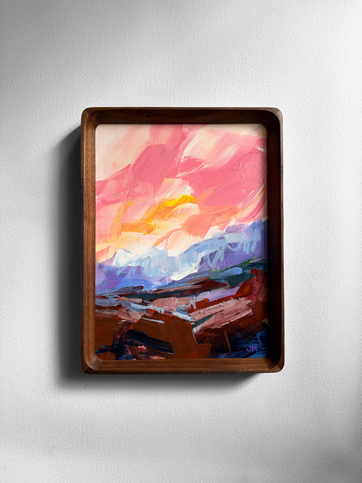 A framed abstract landscape painting featuring vibrant pink, orange, purple, and blue brushstrokes, housed in a dark brown wooden frame and mounted on a light gray textured wall.