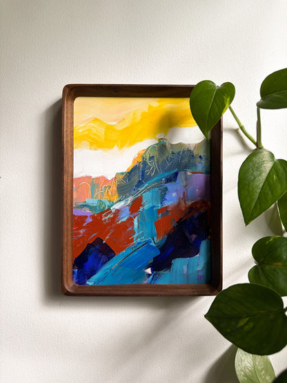 A framed abstract landscape painting with bold brushstrokes in yellow, blue, red, purple, and black, displayed on a white wall next to a green houseplant.