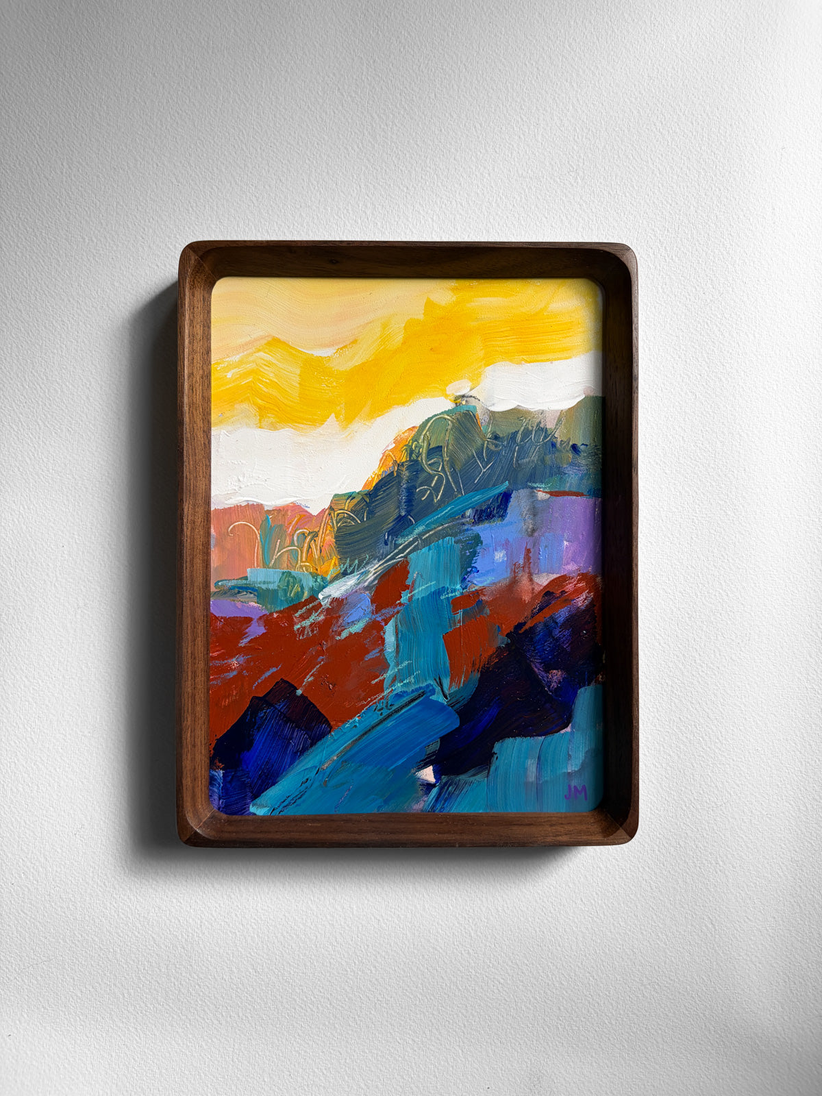 A framed abstract landscape painting with bold brushstrokes in yellow, blue, red, green, and purple, set in a dark brown wooden frame and mounted on a light gray wall.