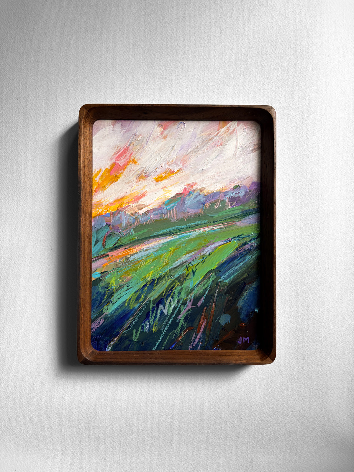 A framed abstract landscape painting featuring vibrant green fields under a colorful sky with orange, pink, and blue hues, housed in a dark brown wooden frame and mounted on a light gray wall.