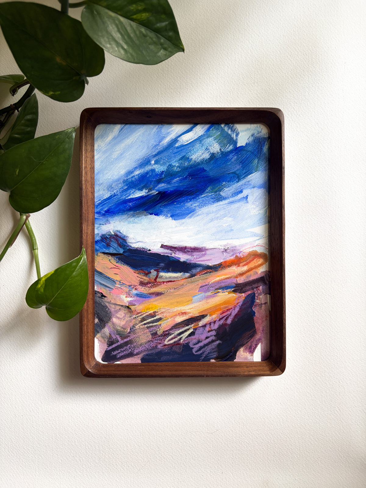 A framed abstract landscape painting with a dark brown wooden frame, hanging on a white wall next to green plant leaves.