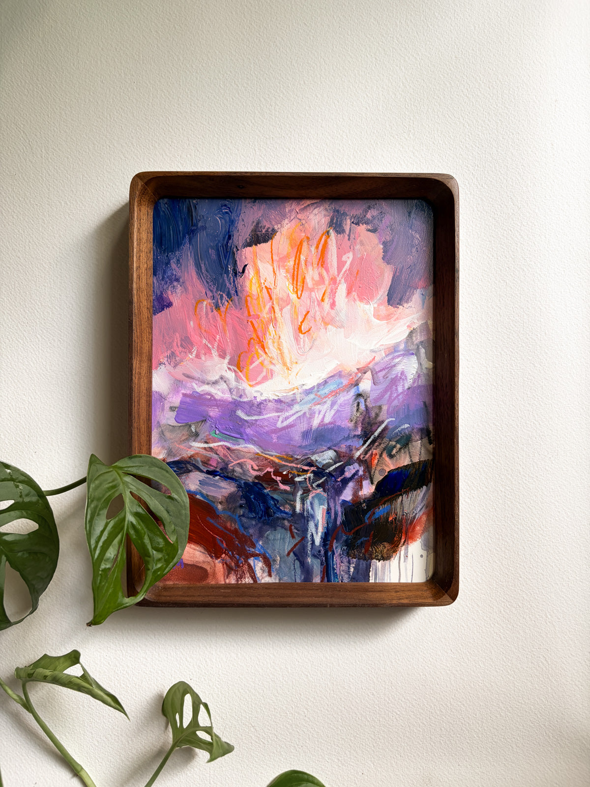 A framed abstract landscape painting with vibrant pink, purple, blue, and orange brushstrokes, displayed on a light beige wall with green plant leaves in the foreground.