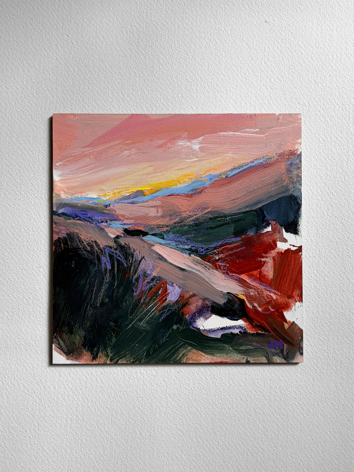 An abstract landscape painting on board, featuring textured brushstrokes of pink, red, yellow, blue, and dark green, displayed on a textured white wall.