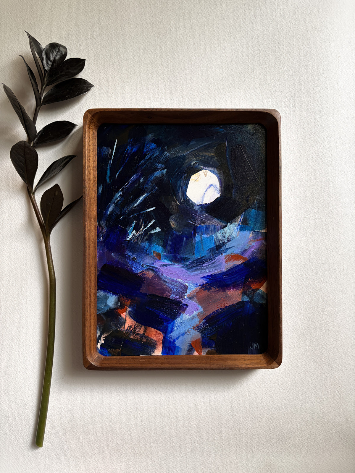 A framed abstract painting featuring a dark night sky with a bright moon, hung on a light beige wall next to a decorative leafy branch.