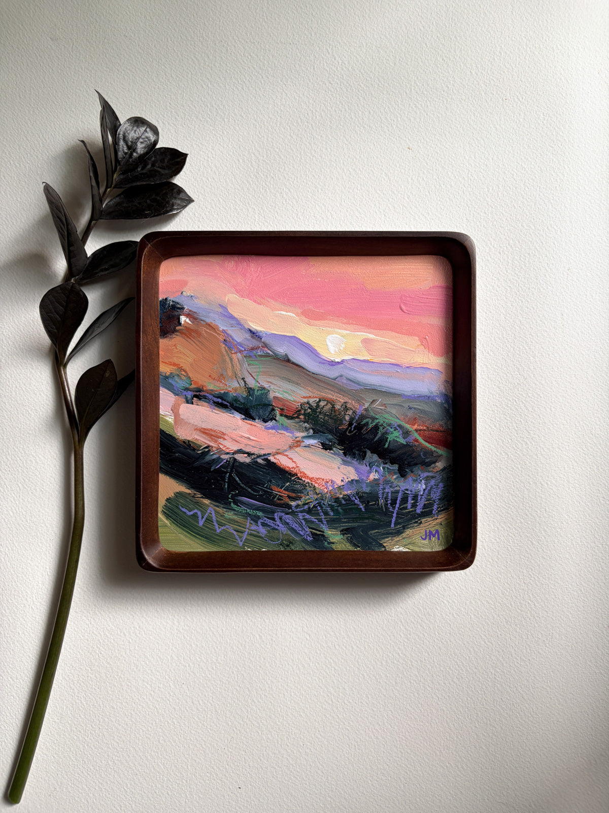 A square framed landscape painting, featuring a pink and orange sunset sky over rolling hills, displayed on a white wall next to a decorative dark green leafy stem.