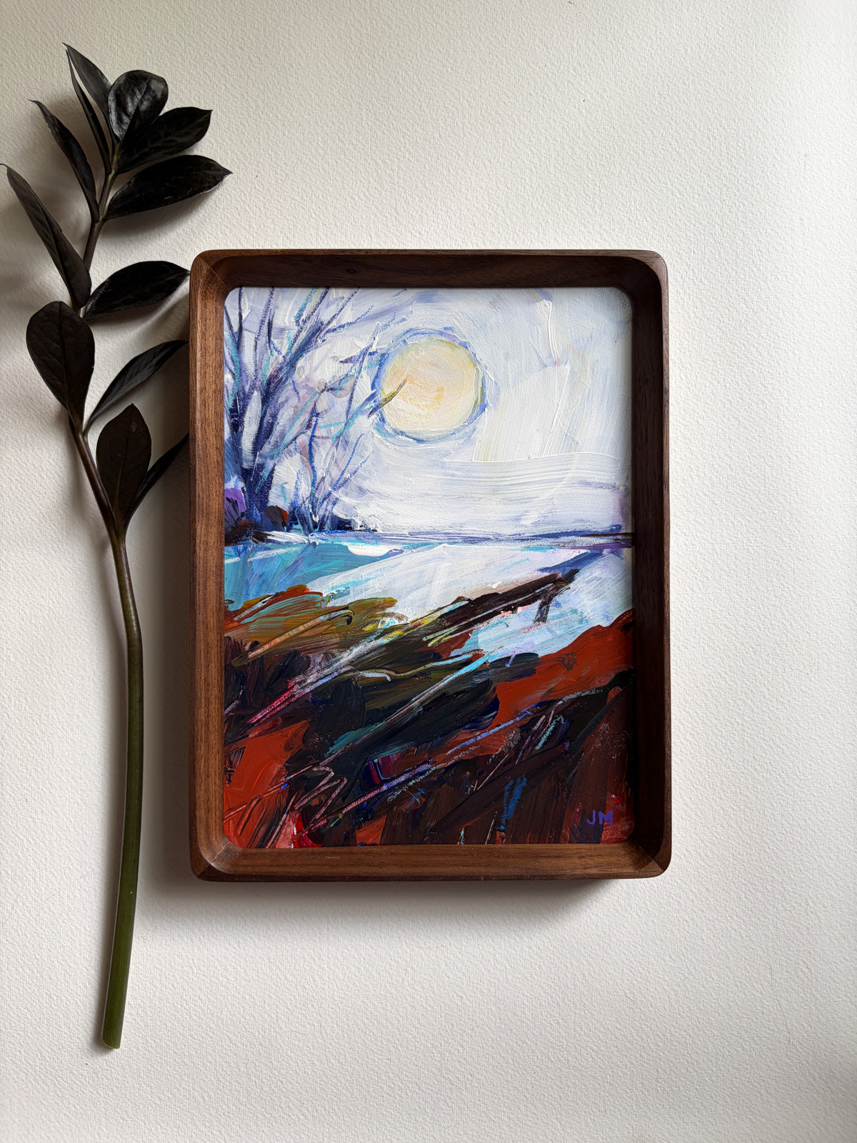A framed landscape painting featuring a moonlit scene with blue water, dark trees, and red-brown foreground brushstrokes, displayed on a light wall next to a dark leafy stem.