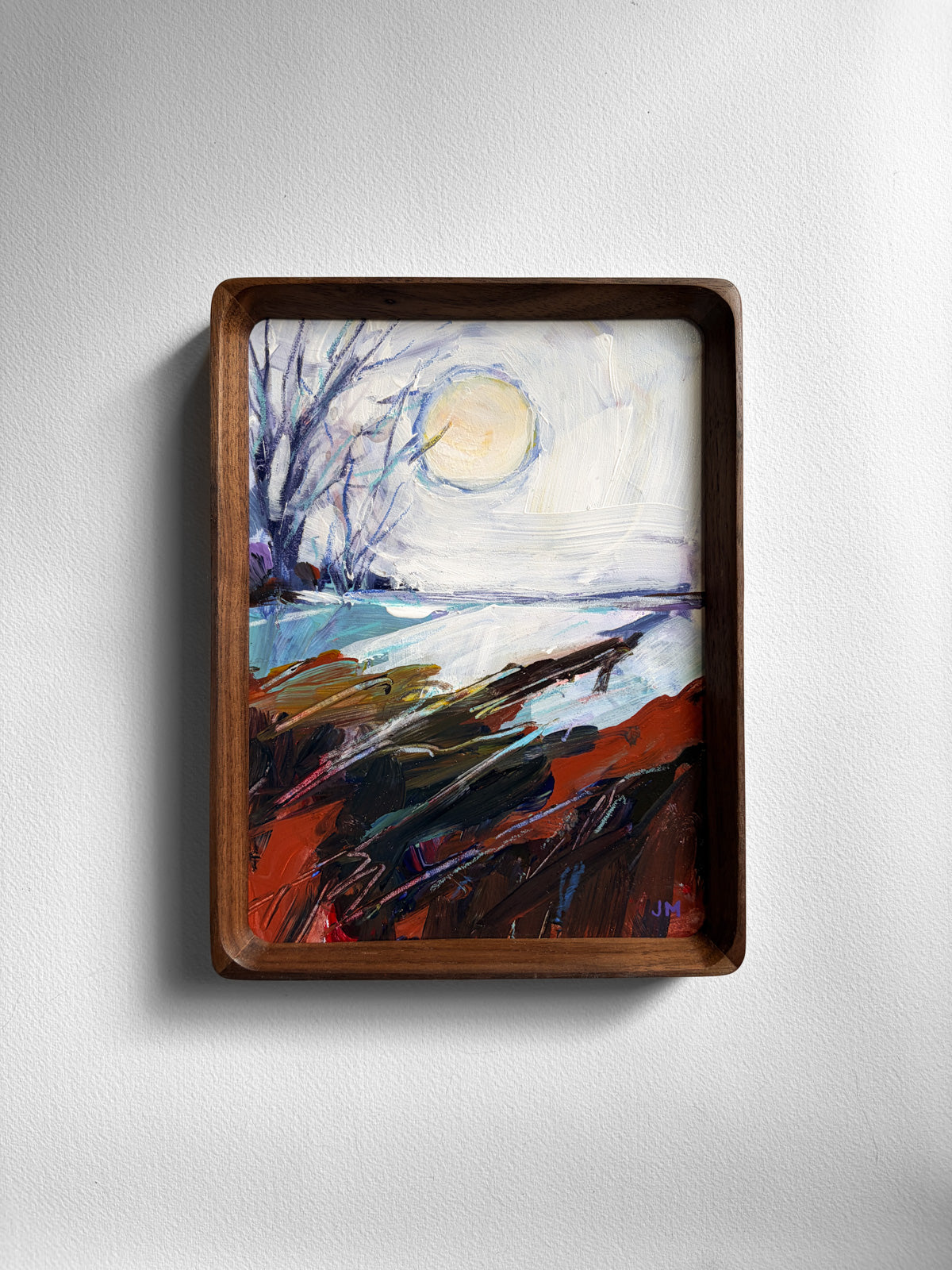 A framed landscape painting hanging on a light gray textured wall. The artwork depicts a moody lakeside scene with bare trees, a pale yellow moon or sun, blue water, and dark reddish-brown foreground brushstrokes.