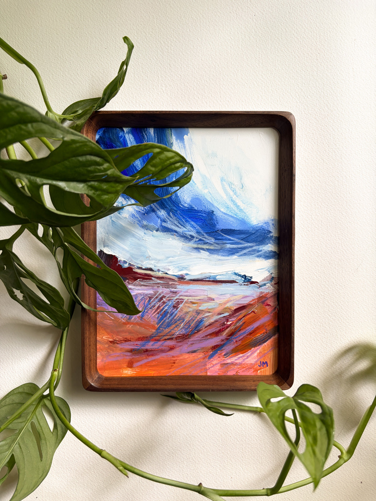 A framed abstract landscape painting with a dark wood frame, featuring blue and white sky-like brushstrokes above a vivid orange, red, and purple ground. Green plant leaves partially cover the left side of the framed artwork against a plain off-white wall.