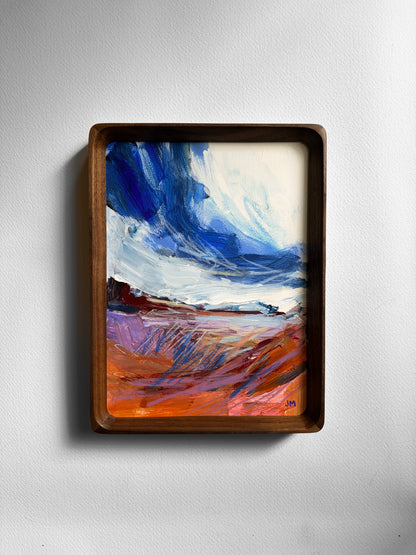 A framed abstract landscape painting featuring a dramatic blue sky and textured orange, purple, and white ground, housed in a dark brown wooden frame and mounted on a light gray wall.