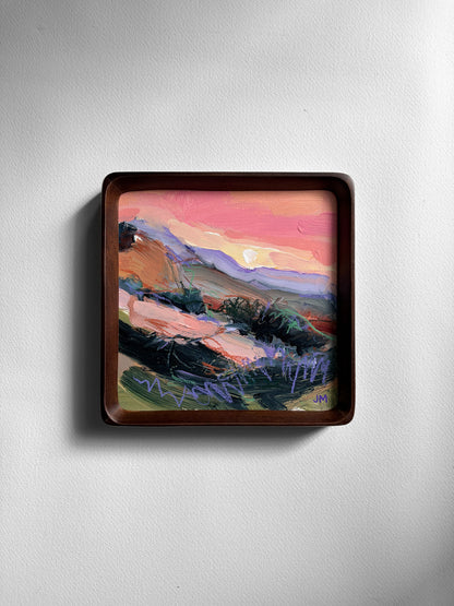 A square framed landscape painting featuring a colorful sunset scene with pink, orange, purple, and green brushstrokes, mounted on a light gray wall.