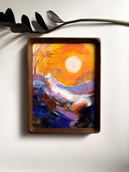 A framed abstract landscape painting featuring a bright yellow and orange sun over purple and blue mountains, displayed on a white wall with a decorative black branch above it.