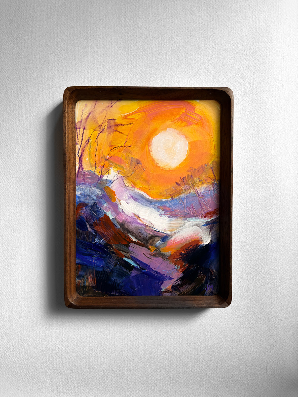 A framed abstract landscape painting featuring a bright yellow and orange sun over purple, blue, and white mountain-like forms, displayed in a dark brown wooden frame against a light gray wall.