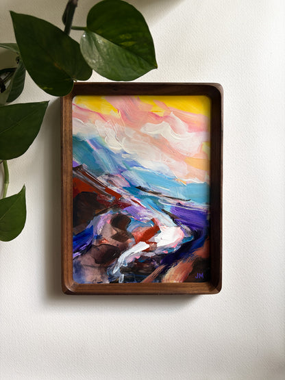 A framed abstract landscape painting with vibrant brushstrokes of blue, purple, pink, yellow, and white, housed in a dark brown wooden frame and mounted on a white wall with green plant leaves hanging in the upper left.
