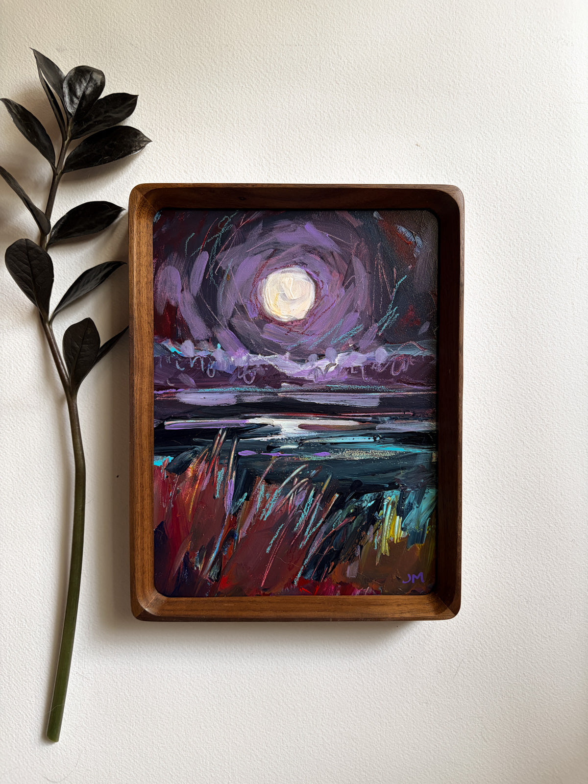 A framed painting of a moonlit purple sky over a dark landscape, displayed on a light wall next to a decorative leafy stem.