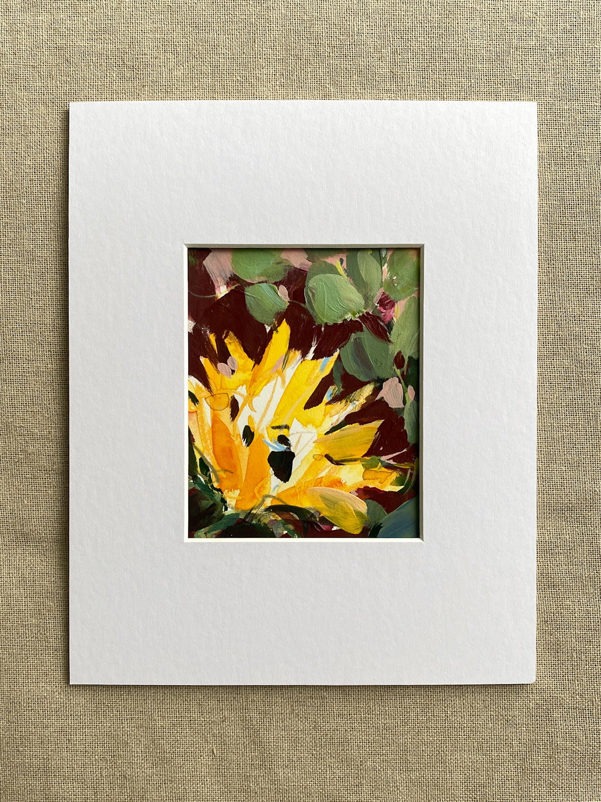 Matted artwork of a yellow sunflower with green leaves on a textured beige background